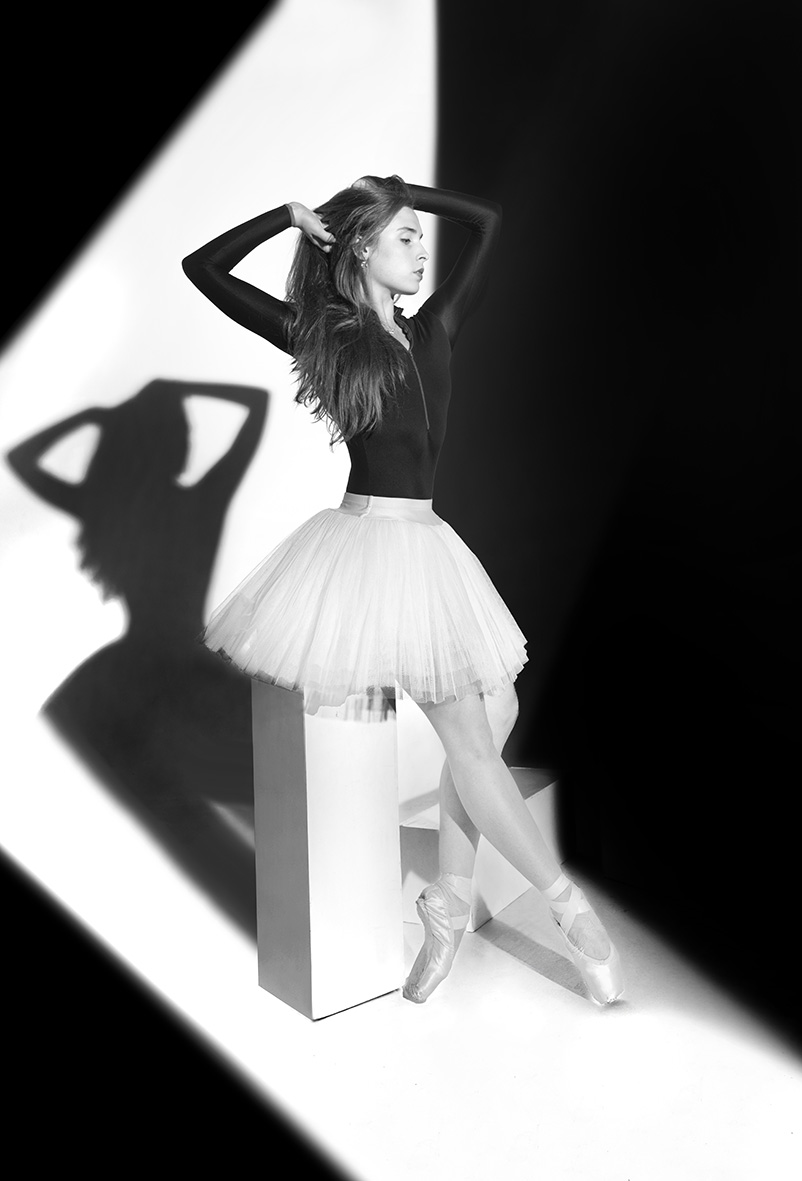 Black-and-white studio photograph of Hannah Martin in a white tutu, framed by geometric light and shadow